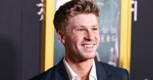 A young man with light brown hair, wearing a dark suit and white shirt, smiles while standing in front of a blurred movie poster at an event.