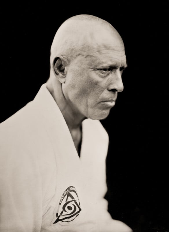 A serious, older bald man in a white martial arts gi with a triangular patch on the chest, shown in profile against a dark background.
