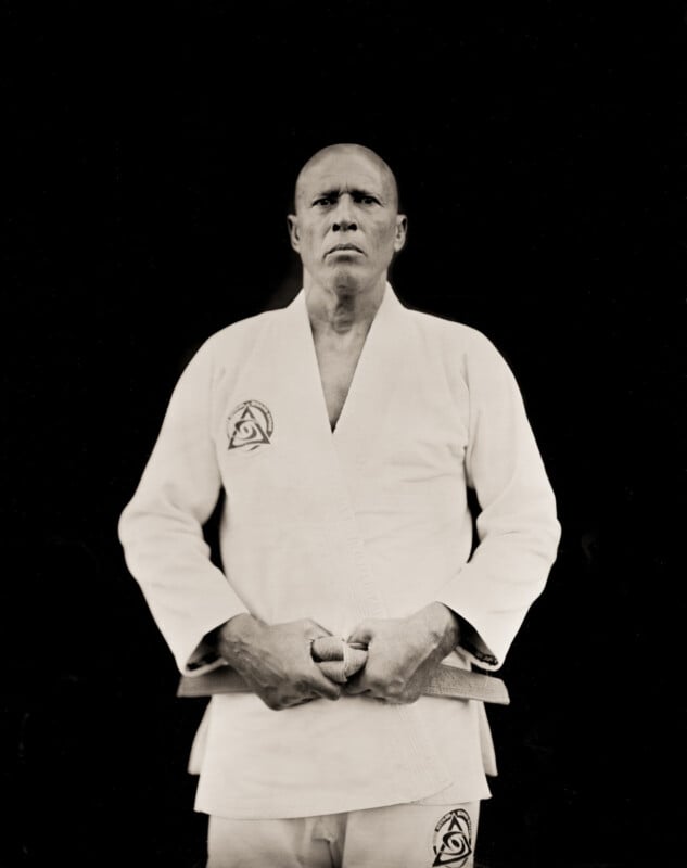 A serious man wearing a white martial arts gi with patches stands against a black background, facing forward with his hands clasped at his waist.