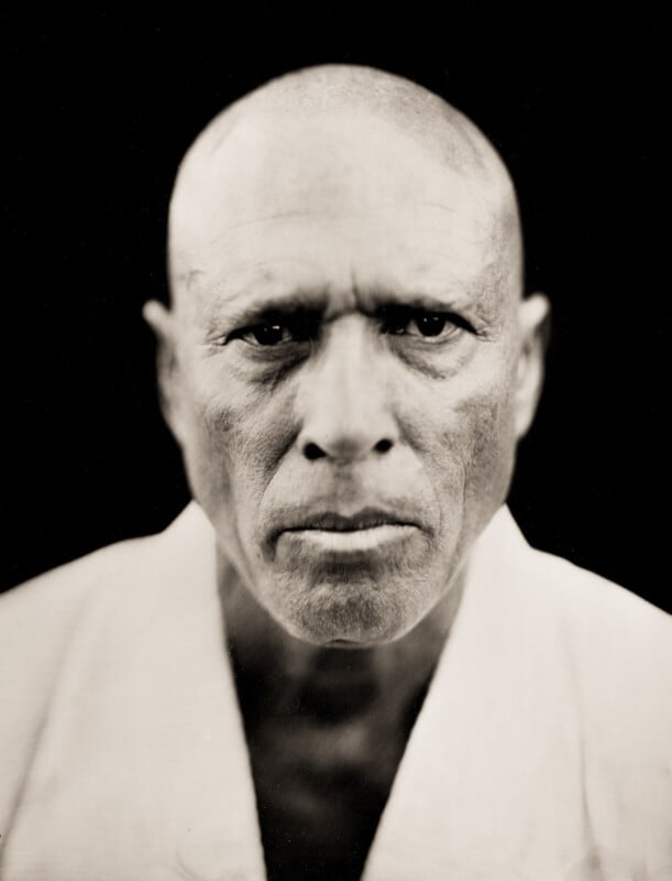 A black-and-white close-up portrait of a serious, bald man wearing a light-colored martial arts gi, looking directly at the camera against a dark background.