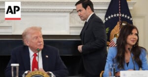 Three people sit at a conference table with microphones. One man in a suit stands in the background. An American flag and presidential seal are visible behind them. The AP logo appears in the upper left corner.