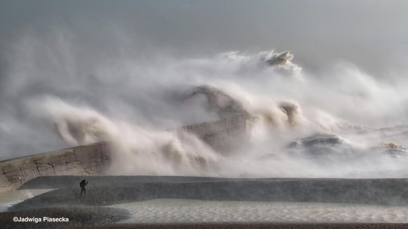 Un hombre se encuentra cerca de un gran malecón mientras enormes olas chocan contra él, creando un espectacular rocío y niebla bajo un cielo nublado.