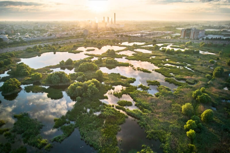Aerial view of wetlands with ponds, green vegetation, and scattered trees at sunrise, with an industrial city skyline and smokestacks in the background under a cloudy sky.