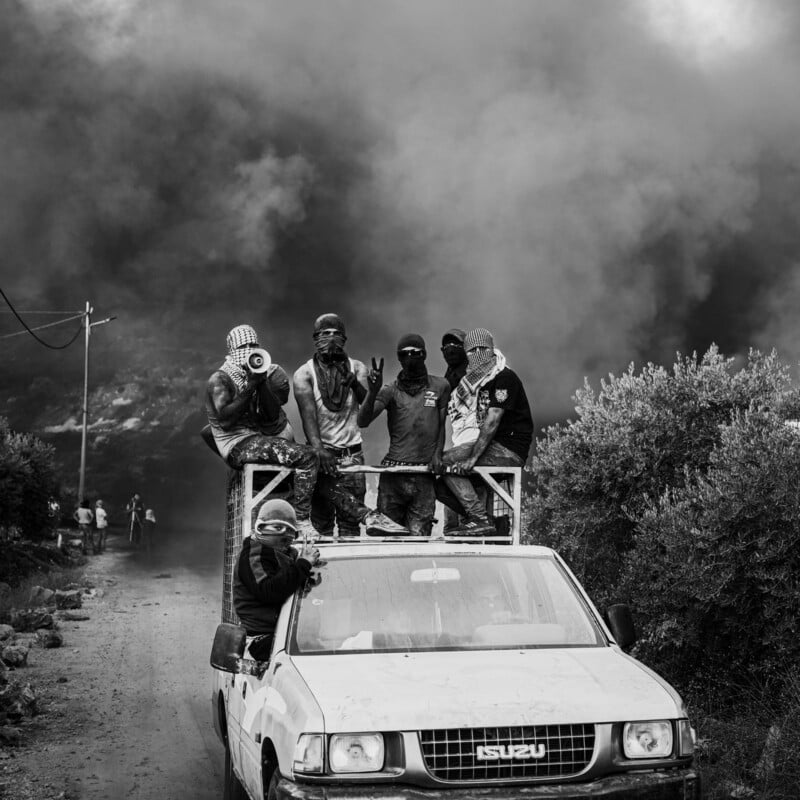 A group of masked people stand and sit on top of a white Isuzu pickup truck, one holding a megaphone, with thick smoke billowing in the background on a dirt road. The scene appears tense and chaotic.