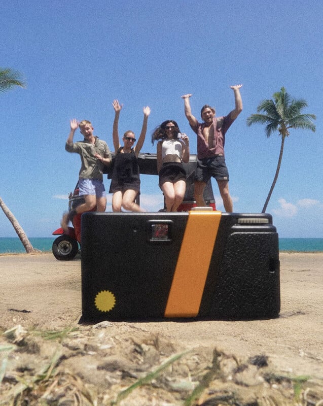 Four friends jump joyfully in front of a red golf cart near the beach, with palm trees, blue sky, and ocean in the background. A vintage camera sits on the ground in the foreground.