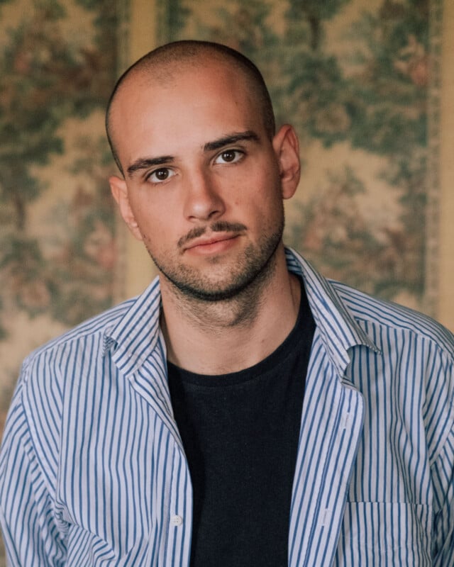 A young man with a shaved head and trimmed beard wears a blue-and-white striped shirt over a black t-shirt, standing in front of a blurred floral patterned background.