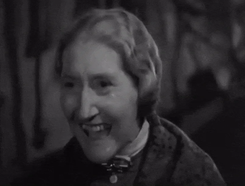 Una fotografía en blanco y negro de una mujer mayor sonriendo. Tiene el pelo corto y ondulado y viste un abrigo estampado con un lazo alrededor del cuello. El fondo está desenfocado.