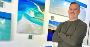 A man with short hair and a beard, wearing a green long-sleeve shirt, stands with arms crossed in front of a wall displaying vibrant landscape photographs, including beaches and ocean scenes.