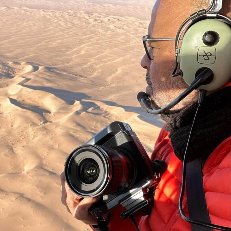 A person wearing a headset and red jacket holds a camera while looking out at a vast sandy desert landscape, possibly from a helicopter or plane. The sunlight casts shadows on the dunes below.