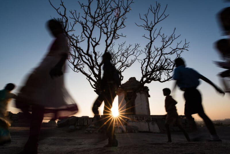 Silhouetted children run past a leafless tree and ancient stone structure at sunset, with the sun’s rays peeking through branches against a clear sky.