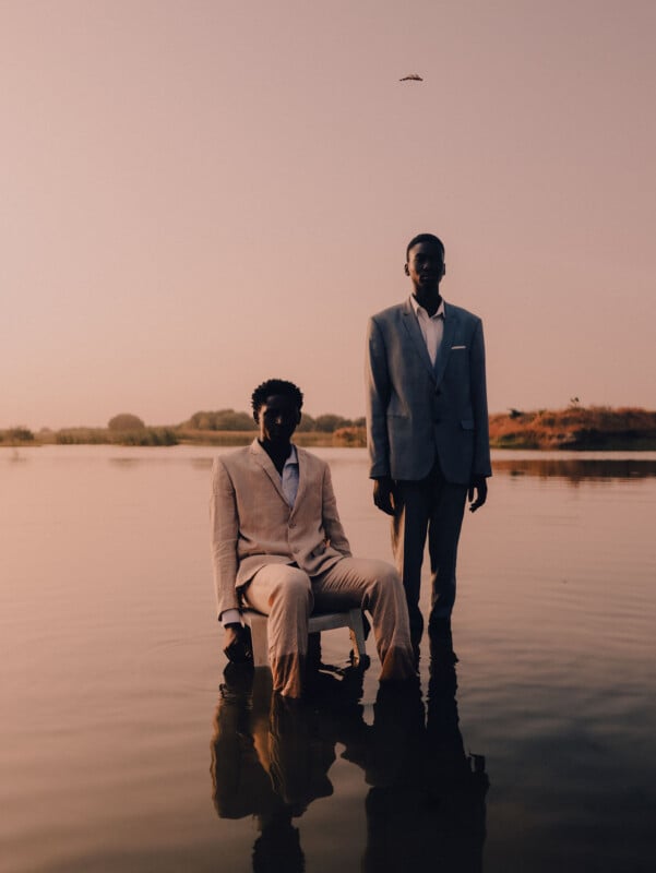 Al atardecer, dos personas trajeadas se sentaron y posaron en aguas poco profundas. El fondo son aguas tranquilas, árboles distantes y un cálido cielo rosa anaranjado. Un objeto pequeño o un pájaro vuela por encima.