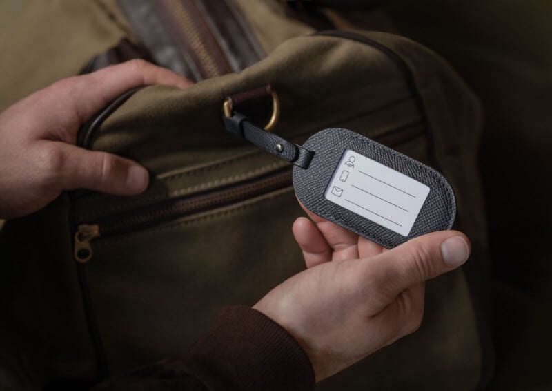 Un hombre sostiene una bolsa de viaje marrón y examina la etiqueta de equipaje gris en blanco pegada a ella. La etiqueta contiene iconos y líneas para datos personales, pero no está completa.