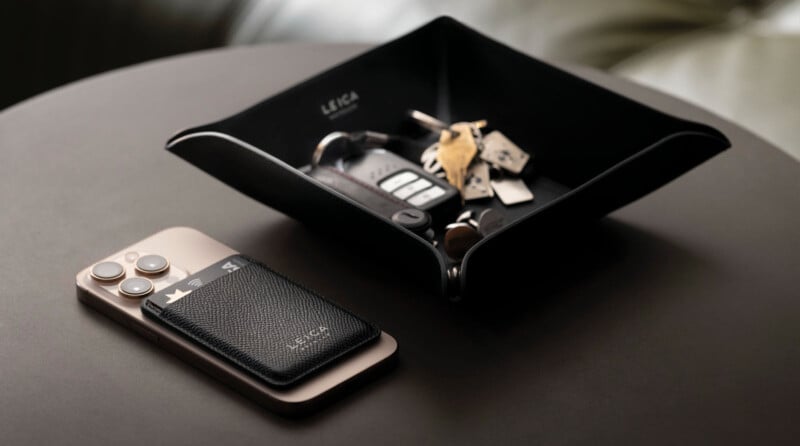 Sobre una mesa redonda oscura hay un smartphone con una funda de cuero negro y una pequeña bandeja con las llaves del coche y otras llaves. Hay etiquetas en el palet. "Leica." La iluminación del lugar es suave, creando un ambiente elegante.