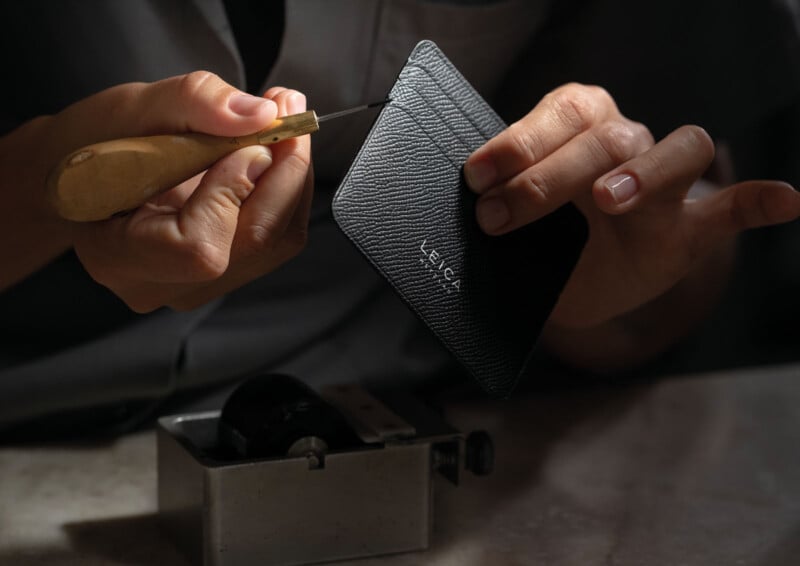 Un hombre trabaja con una herramienta en un tarjetero de cuero con textura negra "Leica" Tiene un patrón en relieve y junto a él hay un pequeño dispositivo de metal sobre la mesa.