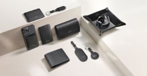 A variety of black leather accessories, including wallets, card holders, keychains, and a valet tray, are neatly arranged on a light beige surface and steps. Some items are embossed with “LUCRIN GENEVA” in white.