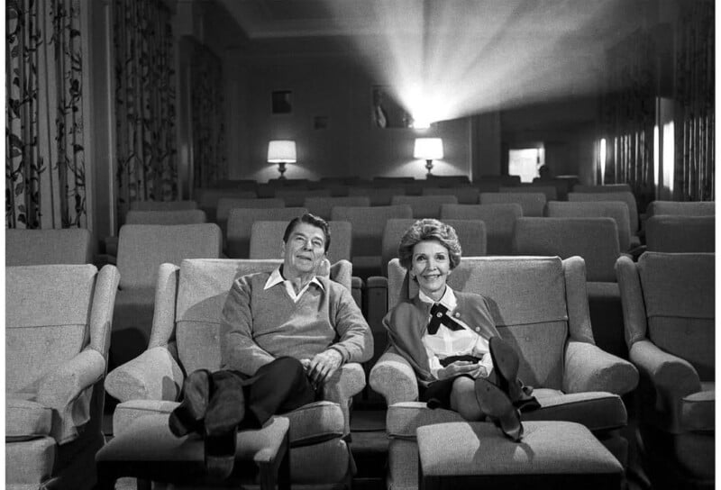 A man and a woman sit together in the front row of an empty theater, smiling as light from a film projector beams across the seats. The cozy room has armchairs and soft lighting from table lamps.