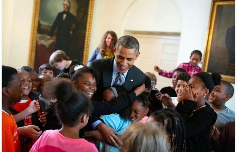 A man in a suit, smiling, hugs and greets a group of excited children in a bright room with large portraits on the walls. The atmosphere is joyful and warm.