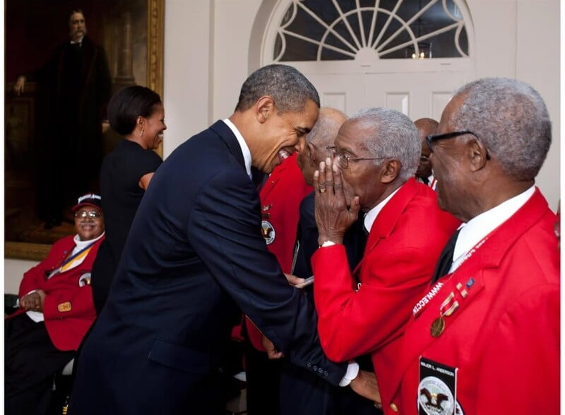 President Barack Obama warmly greets and shakes hands with elderly men in red jackets, likely veterans, while Michelle Obama and others look on and smile in a formal room with a large portrait and arched window.