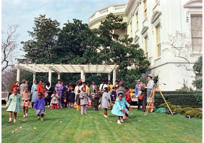 Children and adults gather on the White House lawn near a pergola for an outdoor event, possibly an Easter egg hunt. Some children are holding baskets and searching the grass, while a photographer stands nearby.