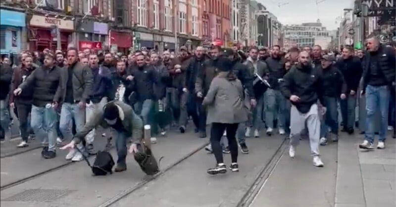 A large group of people, mostly men in dark clothing, are gathered on a city street. Two people are in the foreground, one bending down and the other standing, as the crowd approaches behind them.