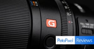 Close-up of a camera lens with a red "G" logo, focusing on detailed markings. The bottom right corner features the Petapixel logo and the word "Reviews" on a blue background.