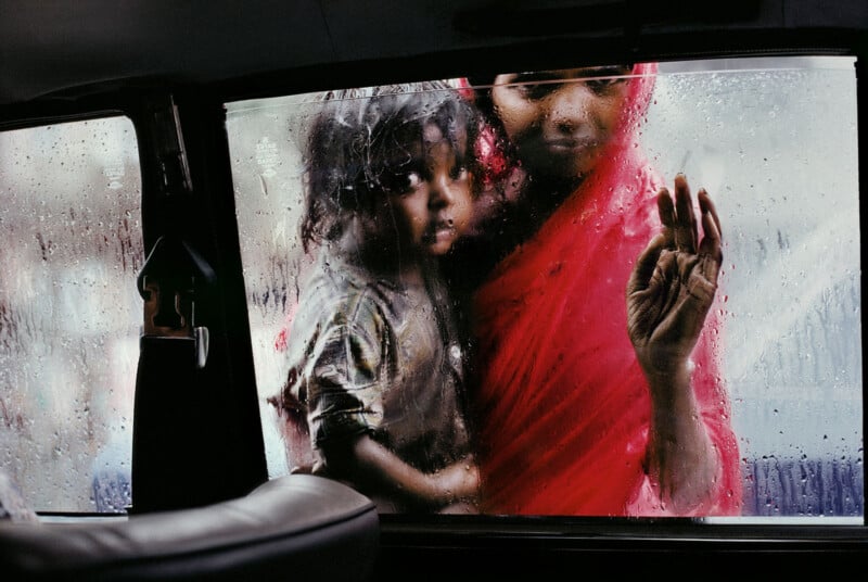A woman in a red sari and a young child stand outside a rain-soaked car window, looking inside. Water droplets blur their faces as the woman rests her hand on the glass.