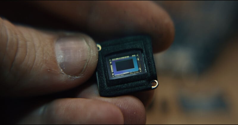 Primer plano de los dedos ligeramente sucios de un hombre que sostienen un pequeño sensor electrónico rectangular o un microchip con contactos metálicos visibles.