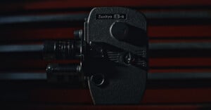 A vintage Sankyo 8-R movie camera is shown close-up against a dark background with red horizontal lines, highlighting its textured body and lens details.