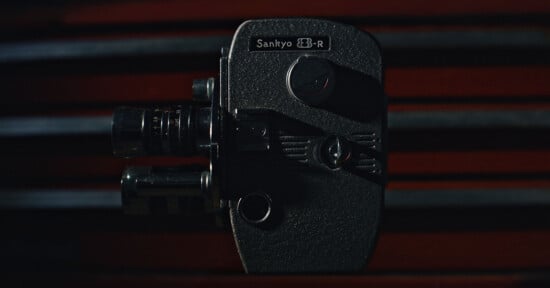 A vintage Sankyo 8-R movie camera is shown close-up against a dark background with red horizontal lines, highlighting its textured body and lens details.