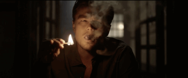 Un hombre está sentado en una habitación con poca luz, se acerca una cerilla encendida a la cara y expulsa humo por la boca, oscureciendo parte de su rostro. La luz de la ventana aparece borrosa en el fondo.