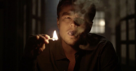 A man in a dimly lit room holds a lit match close to his face while smoking, with smoke partially obscuring his features.