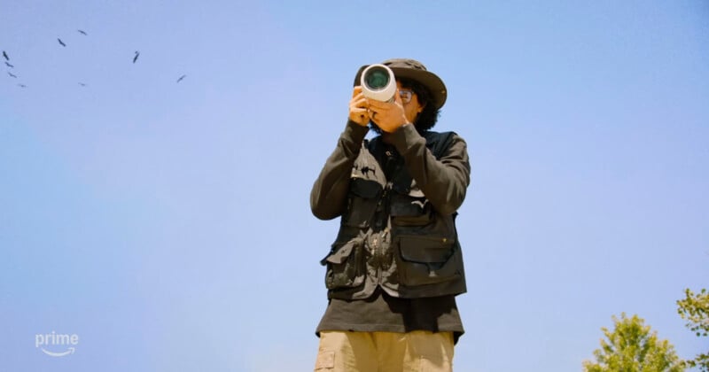 A person wearing a hat and dark vest points a camera with a large lens toward the viewer under a clear blue sky, with birds flying in the distance and greenery in the lower right corner.