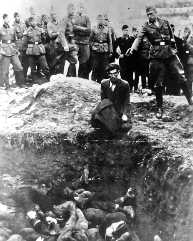 A black-and-white historical photo showing armed soldiers watching as a man kneels at the edge of a mass grave filled with bodies. One soldier points while others observe in the background.