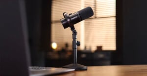 A black Maono microphone on a desk stand sits in focus, with a blurred laptop in the foreground and a softly lit room with blinds in the background.