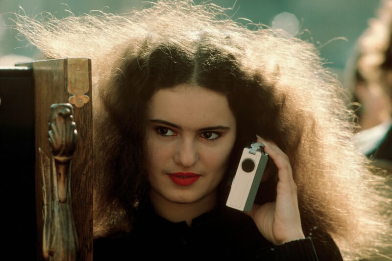 A woman with voluminous, frizzy hair and bright red lipstick holds a vintage portable cassette player to her ear. She stands outdoors, partially next to a wooden object with a carved hand detail.