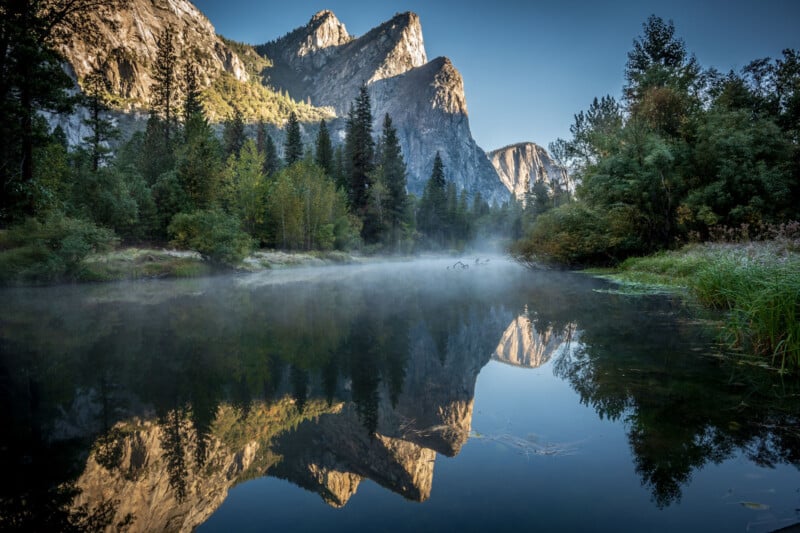 A calm river reflects towering rocky mountains and pine trees under a clear blue sky, with light mist rising from the water in a lush, green forested valley.