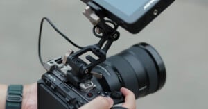 A person holds a Sony camera equipped with a large lens and an external monitor mounted on top, connected by a cable. The camera setup is being adjusted or prepared for use.