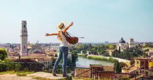 A woman with a hat and backpack stands on a hill with arms outstretched, overlooking a scenic city with historic buildings, a river, and a clear sky.