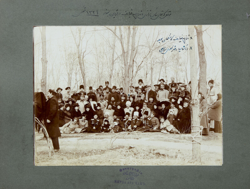 Un nutrido grupo de hombres, mujeres y niños posaron para una fotografía formal al aire libre entre los árboles desnudos. Algunas personas estaban sentadas, otras estaban de pie detrás y algunas estaban paradas a un lado. En la foto hay palabras persas escritas a mano.