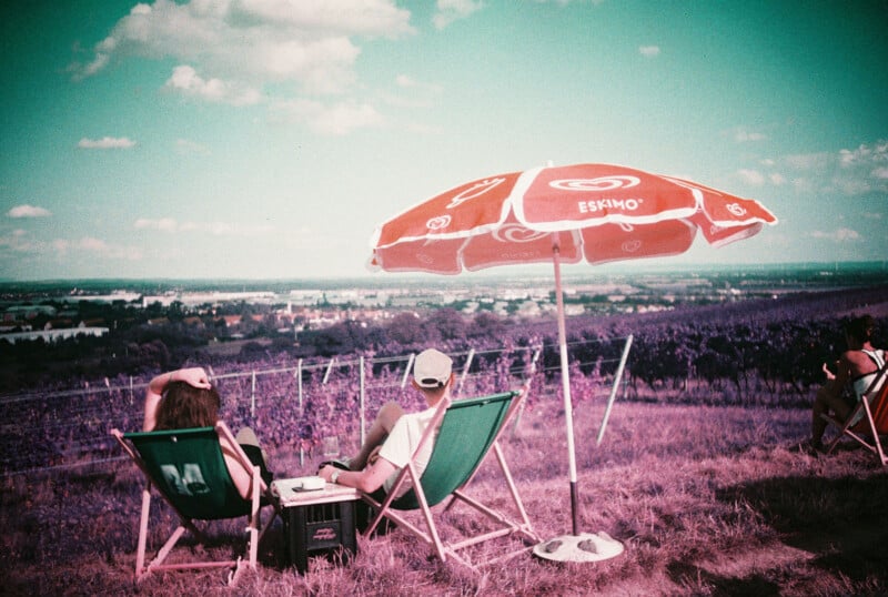 Two people relax in deck chairs under a red Eskimo umbrella on a grassy hill, overlooking a scenic landscape with fields, vineyard rows, and a town in the distance under a partly cloudy sky.