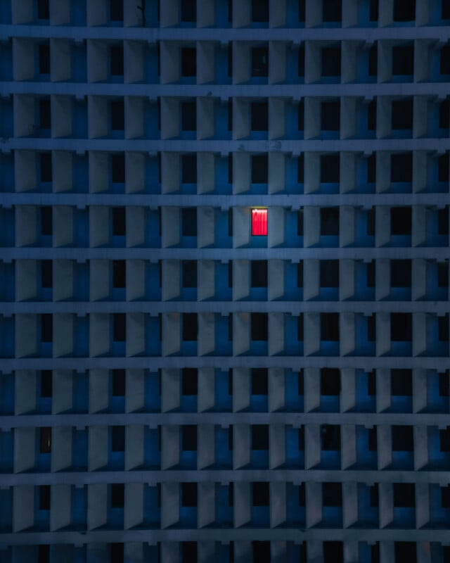 Por la noche, hay una hilera de ventanas azules idénticas en el edificio, una de las cuales está iluminada y cubierta con cortinas de color rojo brillante, lo que la distingue de las demás.