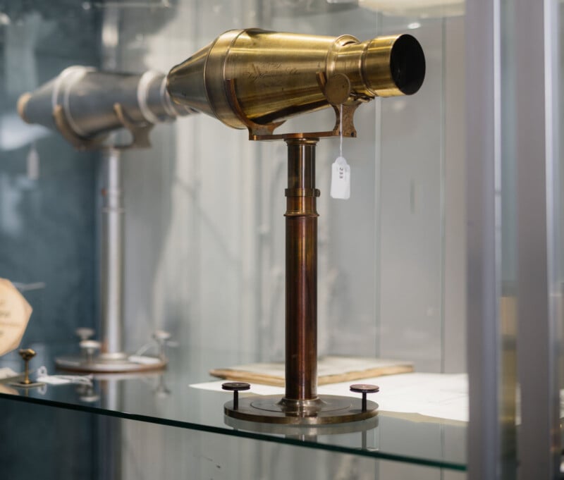 A vintage brass microscope with a cylindrical body and adjustable stand is displayed in a glass case, with another scientific instrument blurred in the background.