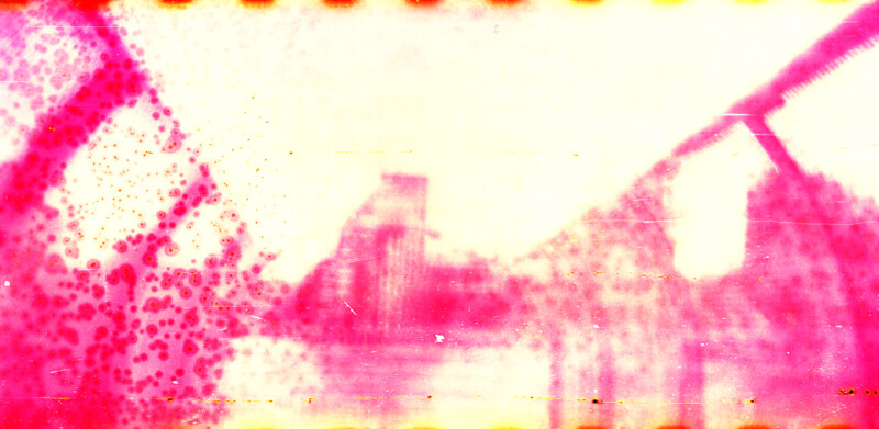 Abstract, faded image with pink and yellow hues. It appears to show a blurry, partially obscured bridge or industrial structure, with irregular dots and marks, giving it a distressed, vintage film effect.