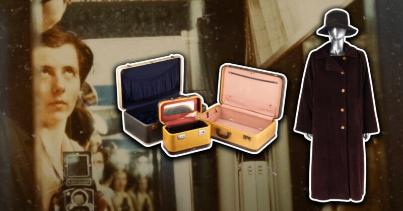 A vintage photo of a woman with a camera is in the background. In the foreground are three vintage suitcases, one open with a red interior, and a long, dark brown coat with gold buttons and a black hat on a mannequin.