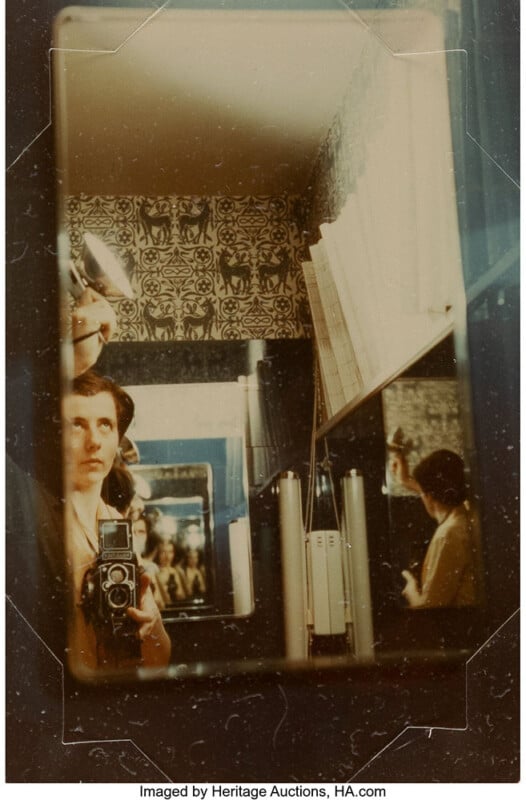 A person takes a self-portrait with a vintage camera in front of a mirror, capturing their reflection along with patterned wallpaper and bookshelves in the background. Multiple reflections are visible in the photograph.