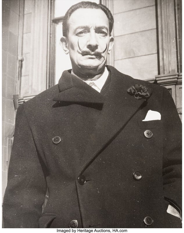 A man with a distinct upward-curled mustache wears a double-breasted coat with a flower pinned on the lapel, standing in front of a building with stone walls.