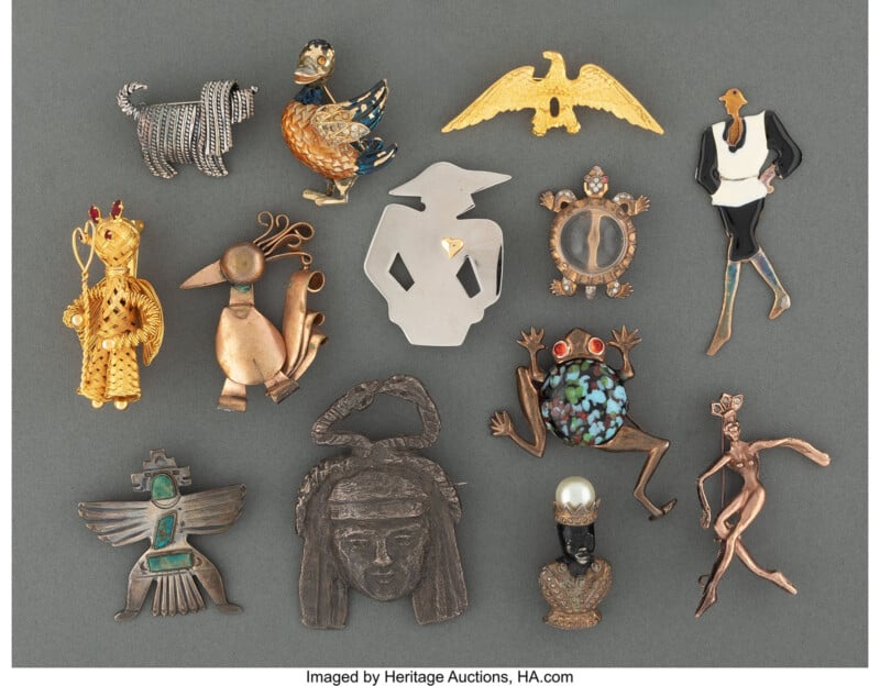 A collection of 14 assorted vintage brooches and pins in various animal, human, and abstract shapes, made from metals and adorned with stones, displayed on a flat gray background.