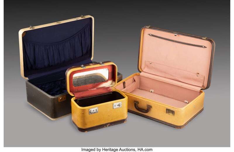 Three vintage suitcases are displayed open: one large black case with a blue interior, one medium tan case with a pink interior, and one small yellow case with a tan interior and built-in mirror.