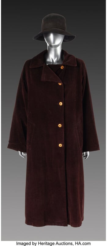 A mannequin wearing a long, dark brown, buttoned coat with large wooden buttons and a matching dark brown wide-brimmed hat, set against a plain gradient background.