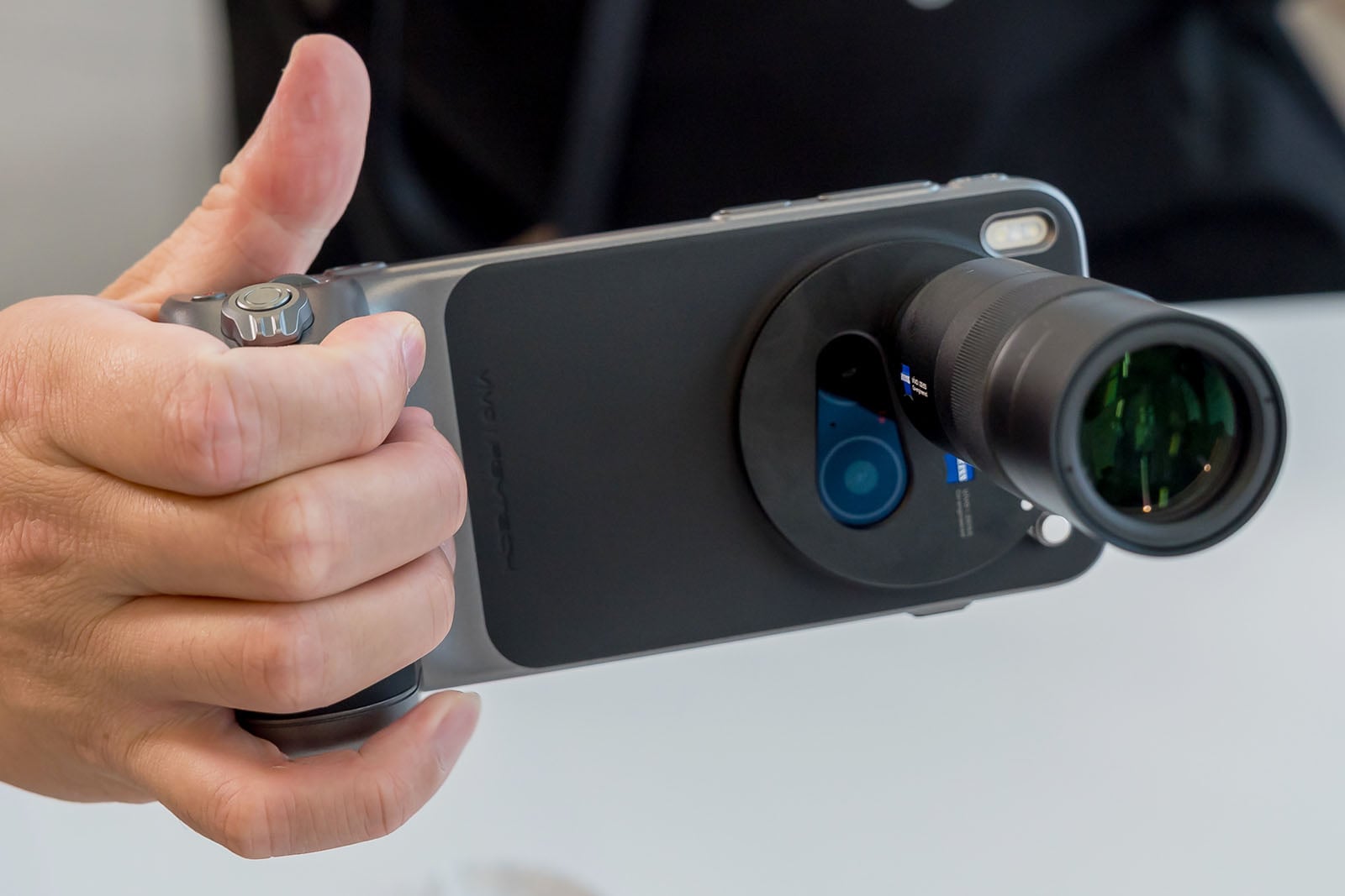 A person holds a smartphone with an attached external camera lens and a grip, enhancing its photography capabilities. The setup is held horizontally, ready for taking photos.